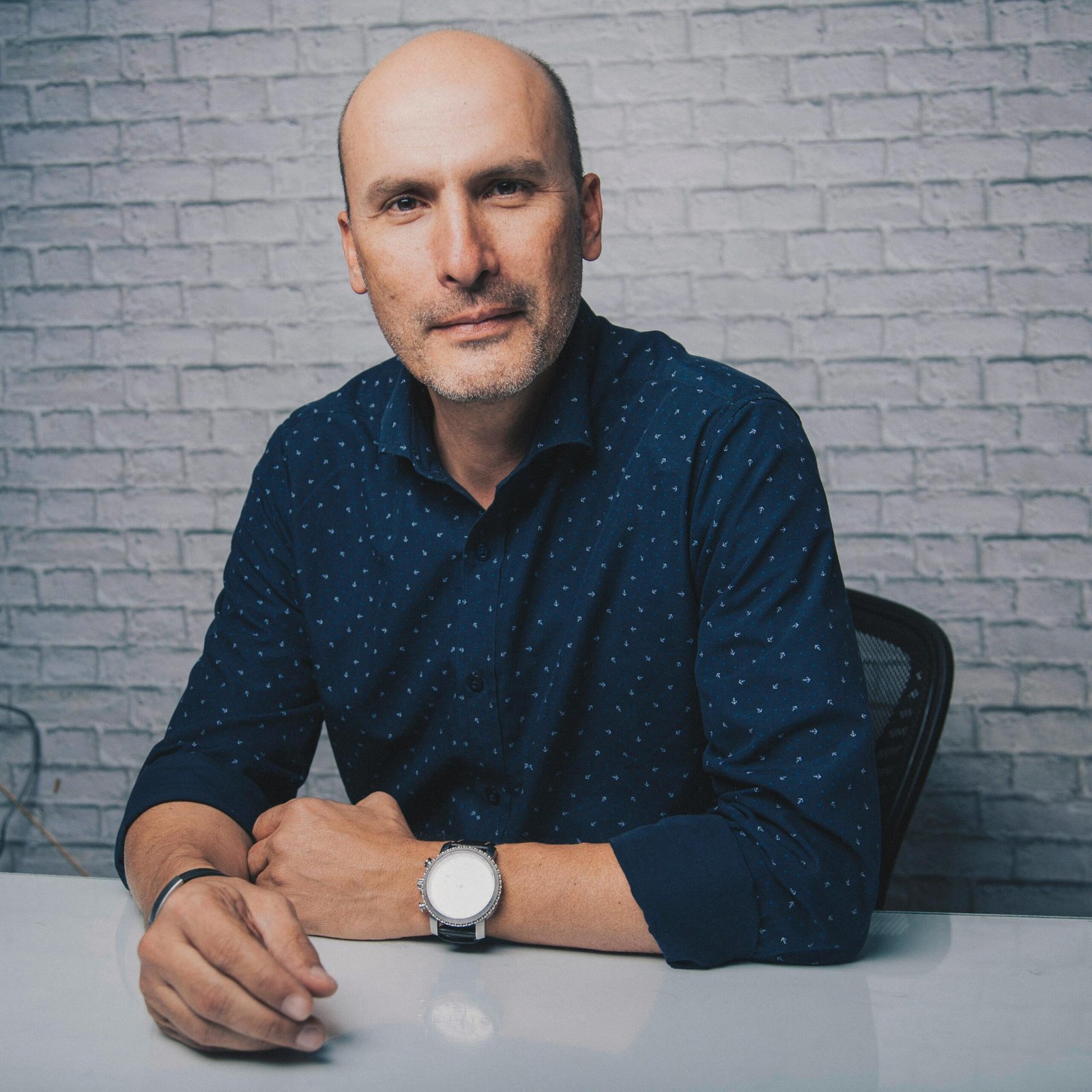 Home A confident middle-aged man sitting in a studio with a brick wall background.